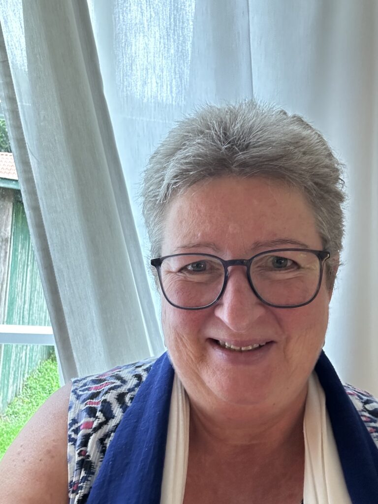 Lächelnde ältere Frau mit kurzen grauen Haaren und Brille, die einen blauen Schal trägt, in der Nähe eines Fensters mit Vorhängen.