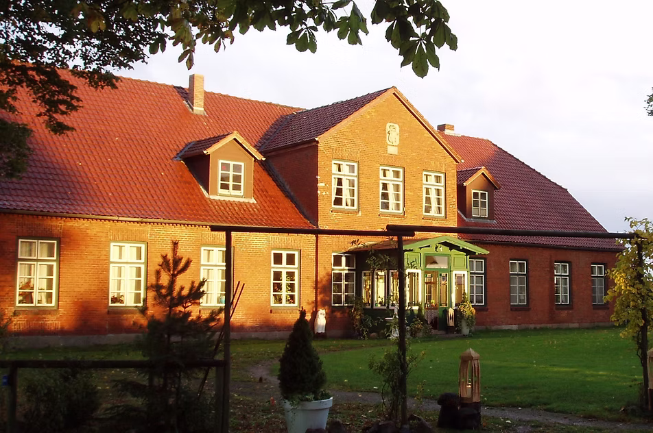 Großes Haus aus rotem Backstein mit vielen Fenstern, rotem Dach und einer grünen Veranda, umgeben von Gras und Bäumen.